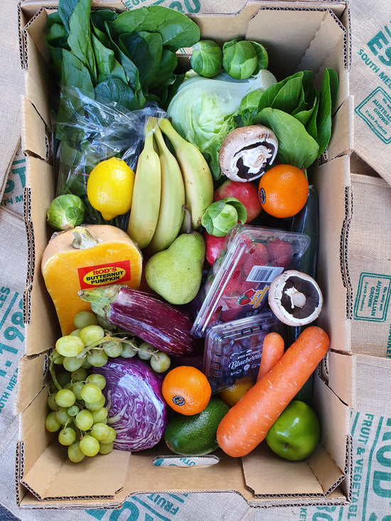 A variety of fresh fruits and vegetables packed in a cardboard box, including bananas, apples, oranges, grapes, carrots, and leafy greens